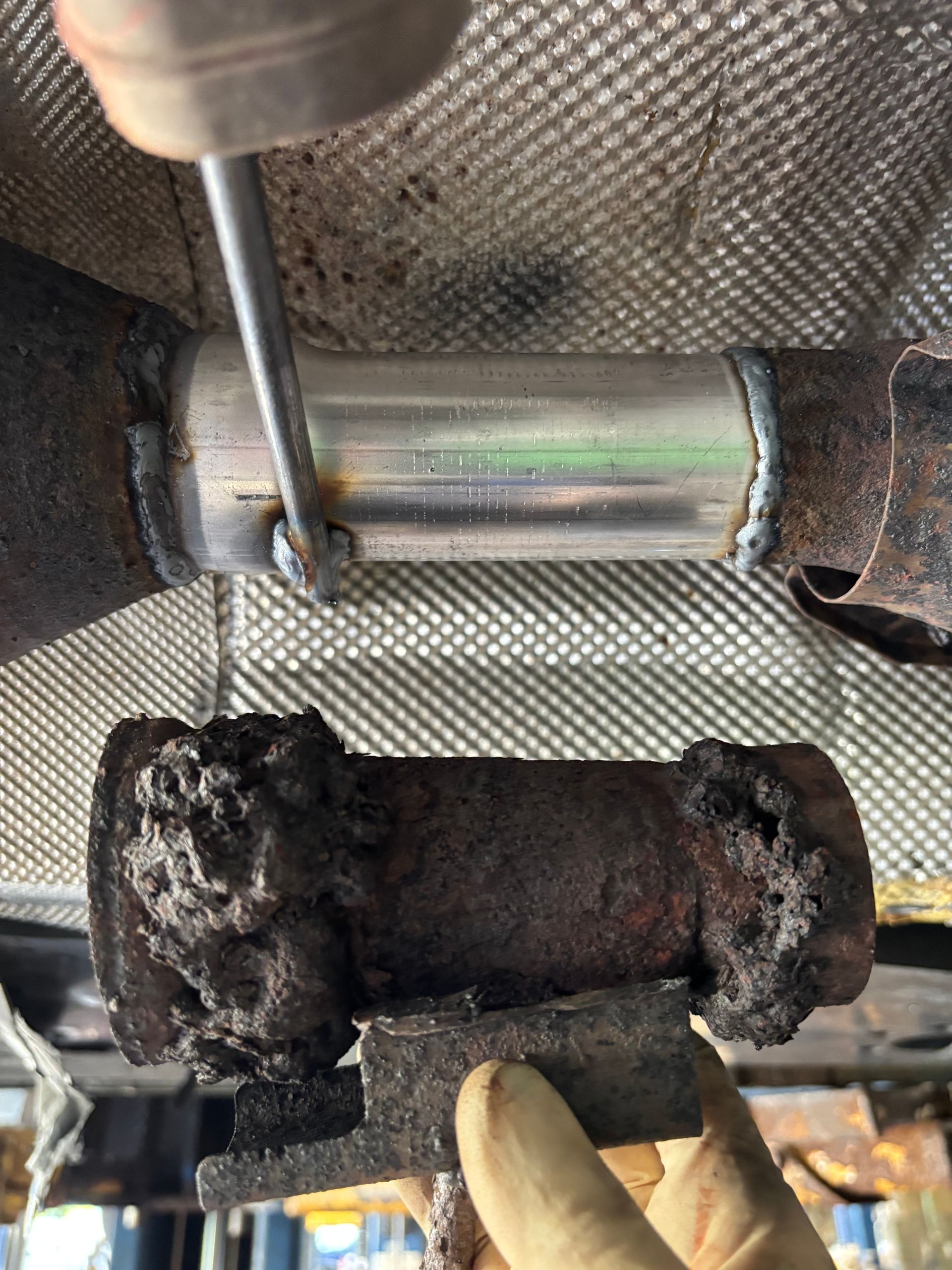 Replacing a rusted exhaust pipe section with a shiny new one; mechanic holding a rusty support.