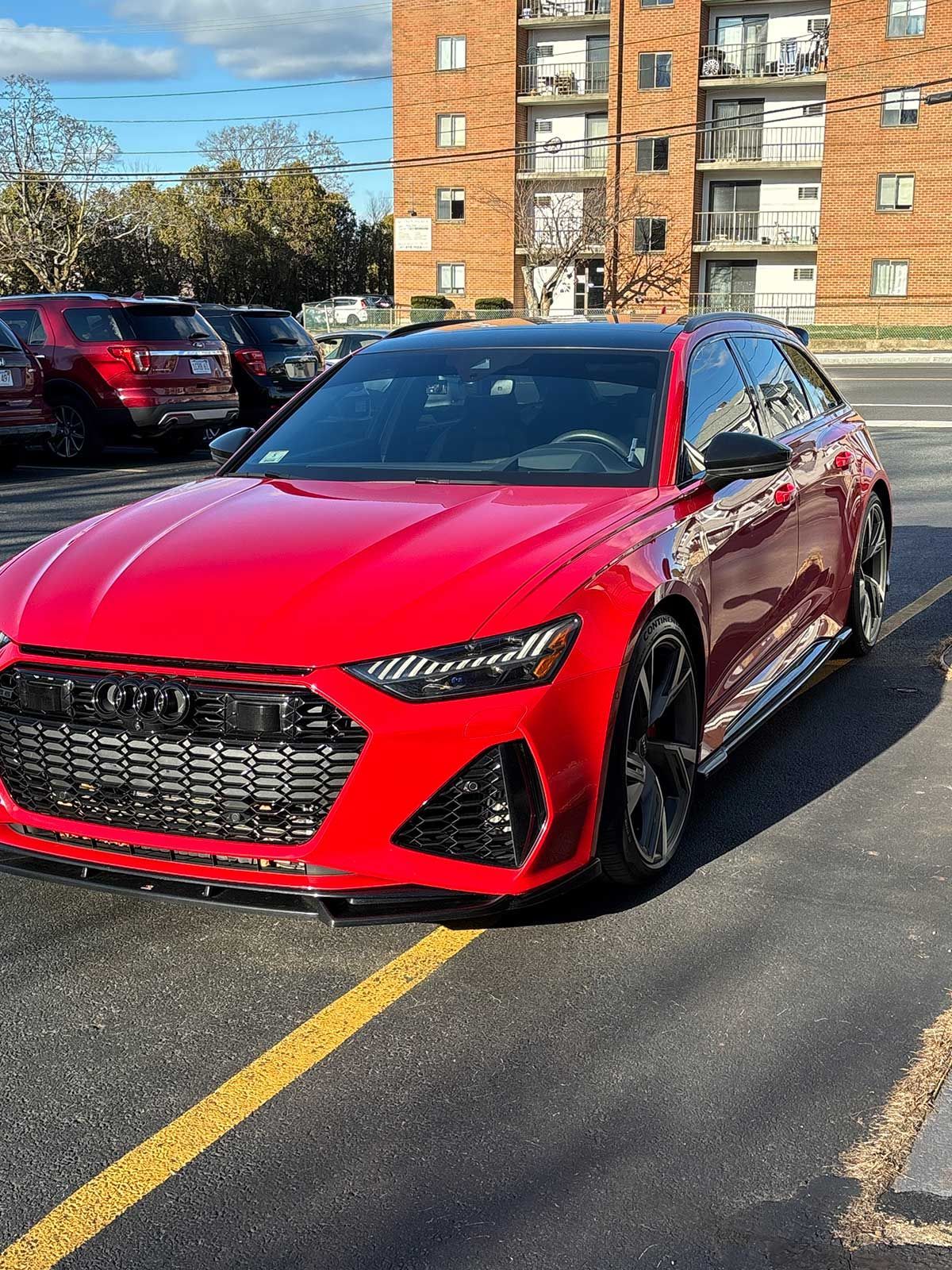 Red Audi RS6 Avant parked in a parking lot. Black accents, tall brick building in background.