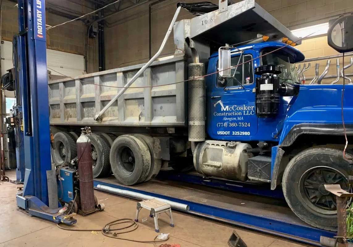 Blue dump truck on a lift in a garage. Welding equipment sits nearby.
