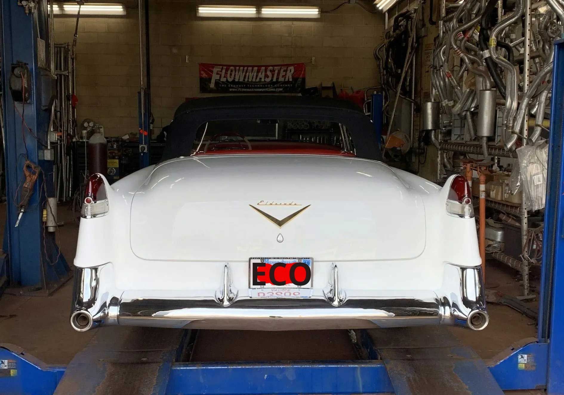 White convertible Cadillac on a lift in a garage, exhaust pipes in the background.