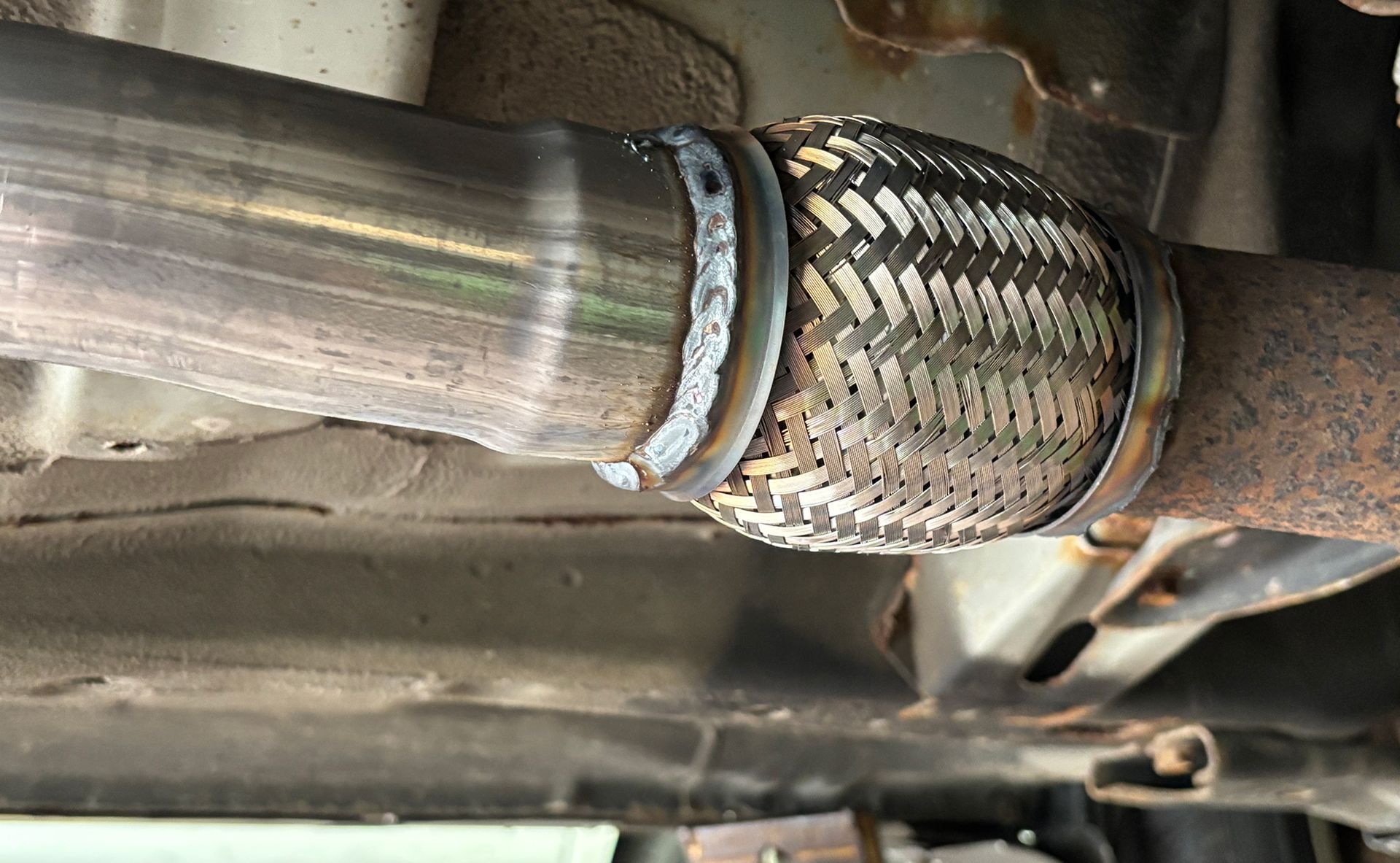 A car's exhaust pipe with a flexible braided section, welded to a rusty metal pipe.