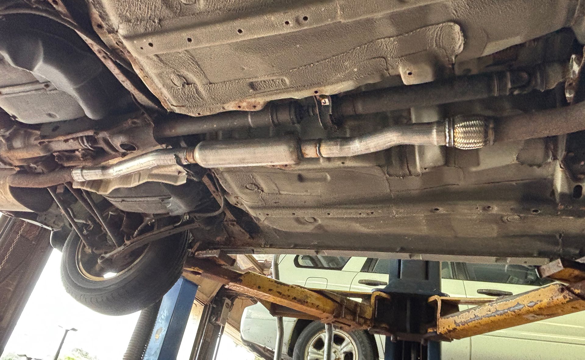 Undercarriage of a vehicle on a lift, showing exhaust system, drive shaft, and rusted metal parts.