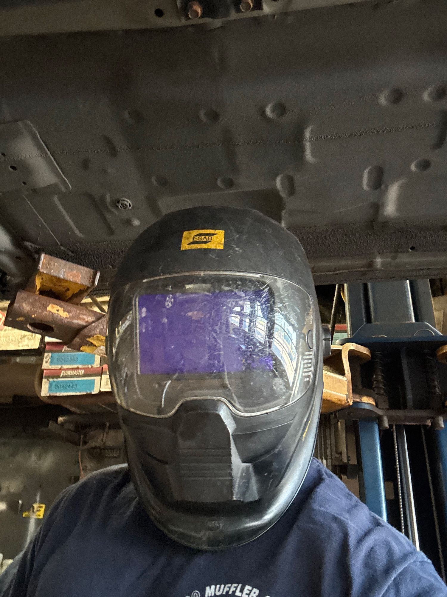Person wearing a black welding helmet, looking up at an underside of a car. In a mechanic shop.