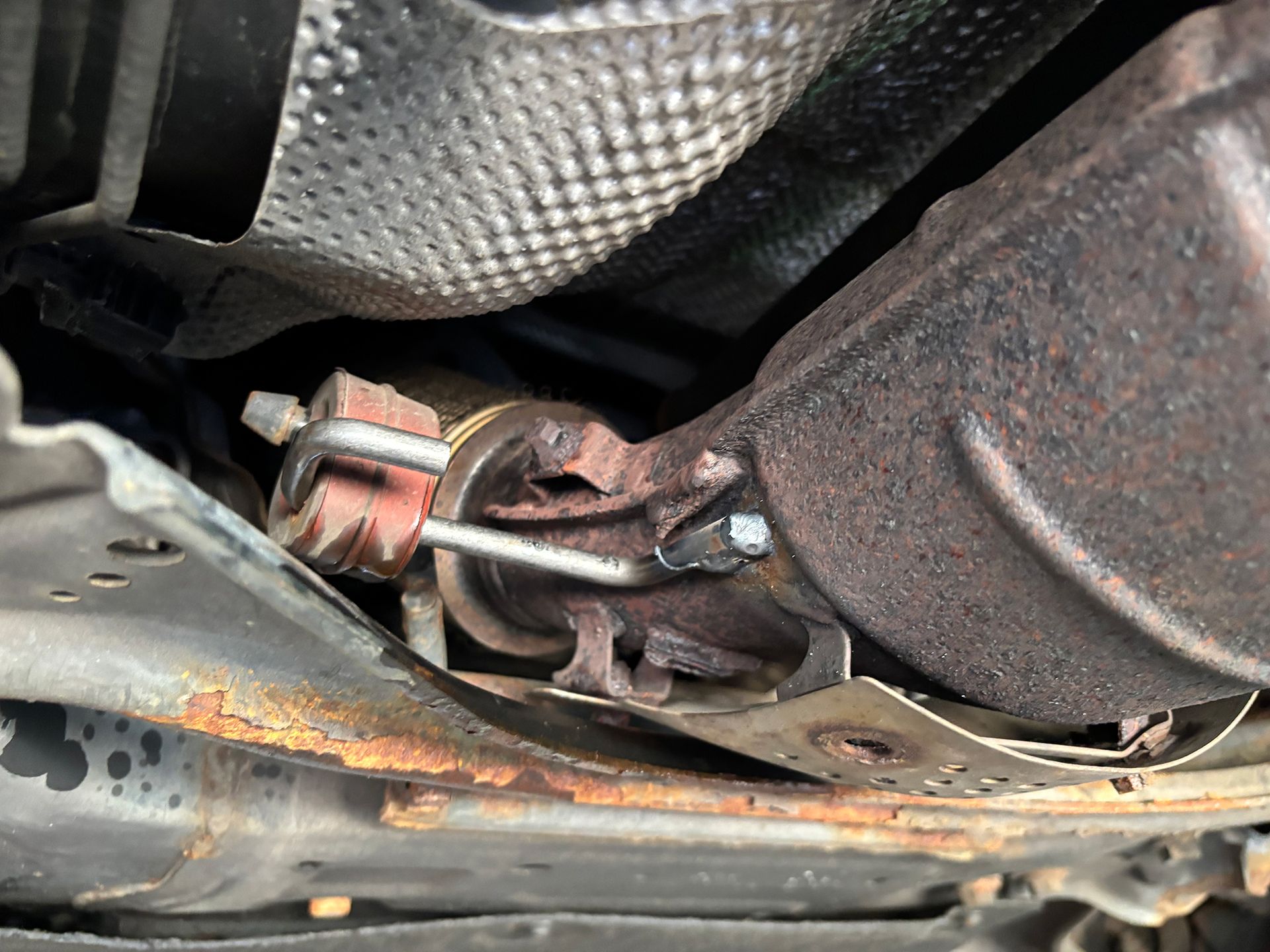 Undercarriage view of a car's exhaust system with a rusty catalytic converter and surrounding components.