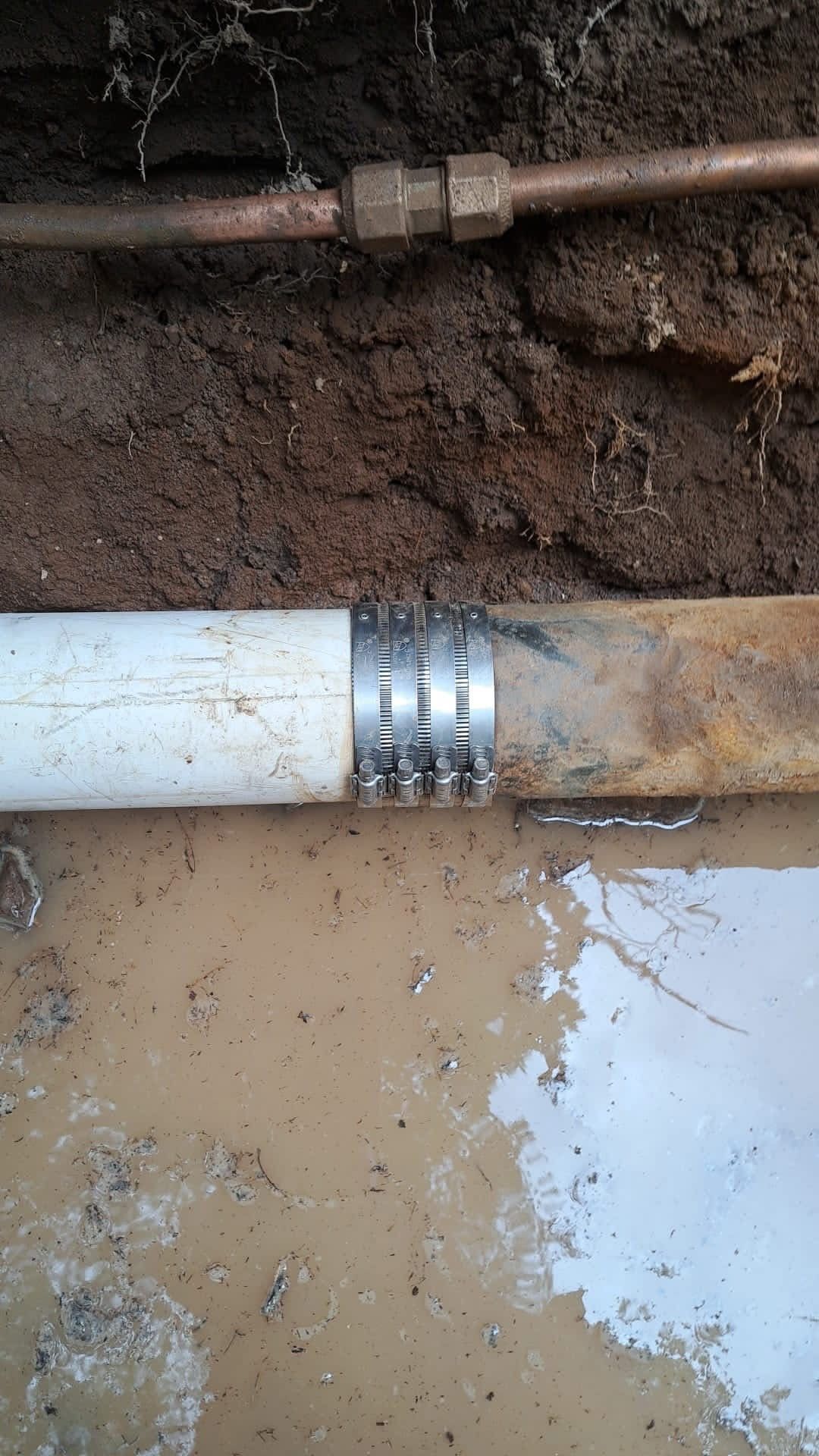 a PVC pipe connected to a corroded pipe with a clamp in muddy ground