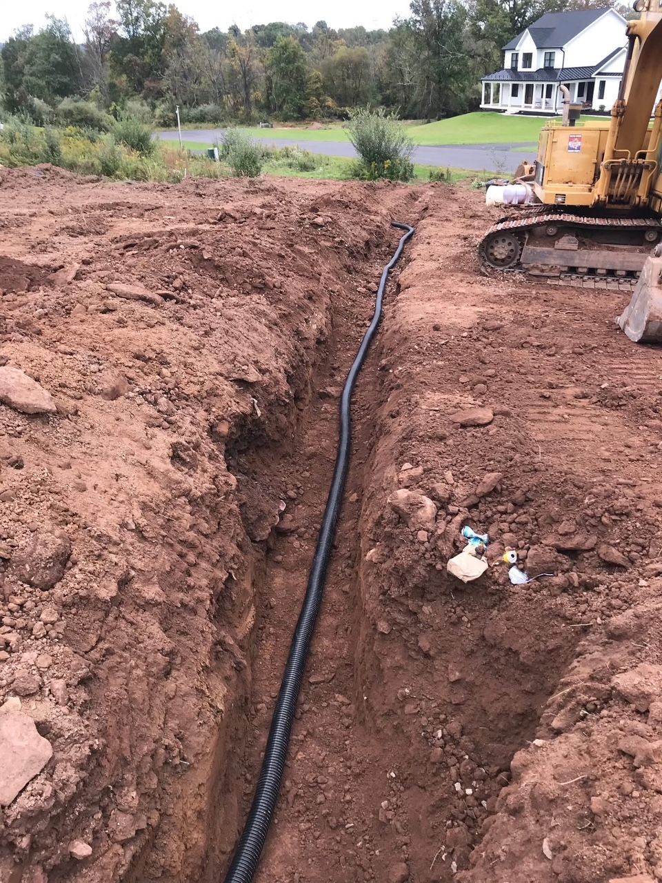trench in dirt with black pipe, excavator, and a house in the background