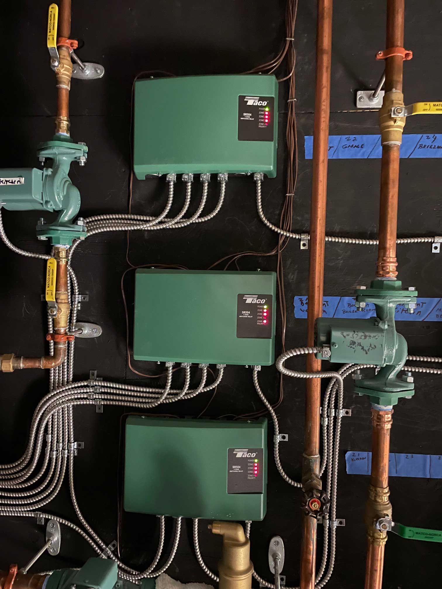 three green control boxes with attached wires, next to copper pipes and pumps on a black wall