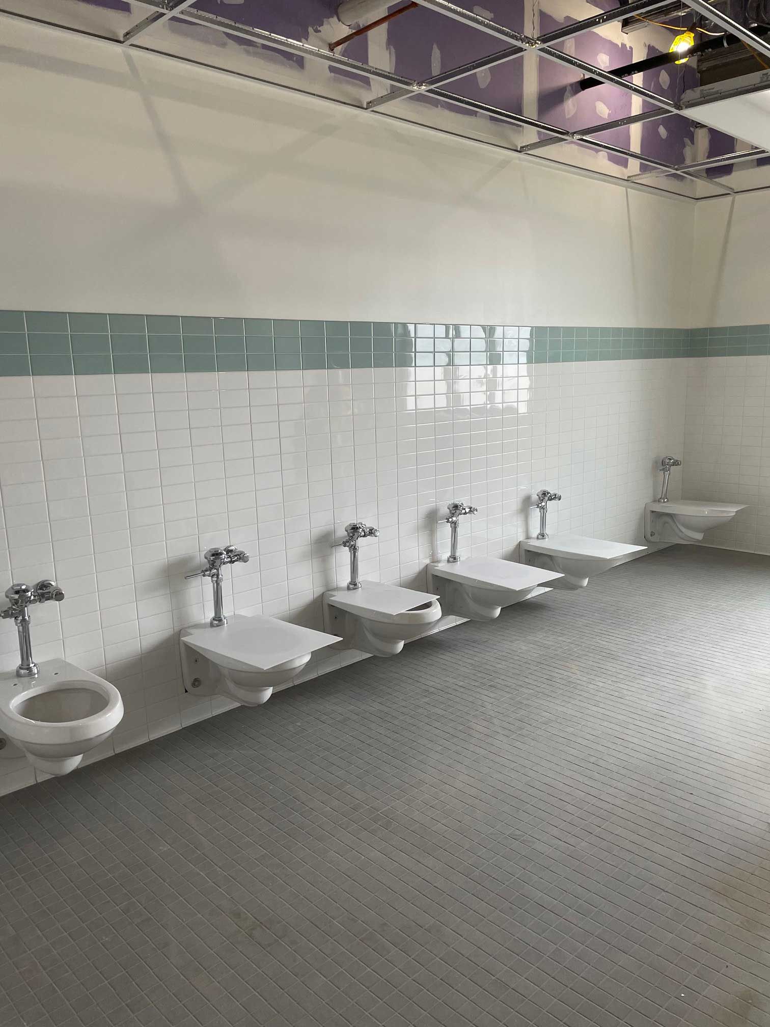a row of white toilets in a public restroom with white and turquoise tiled walls, and a gray floor