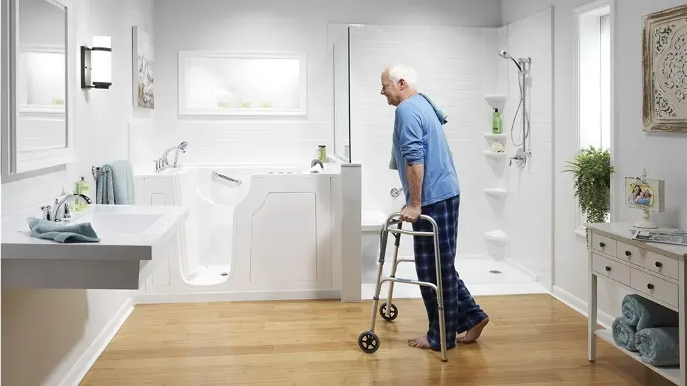 Elderly person with walker in a bright bathroom near a walk-in shower and sink