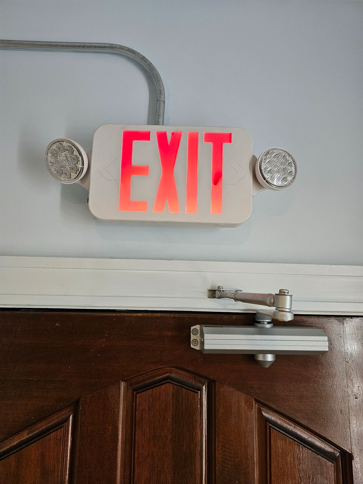 Illuminated red EXIT sign above a wooden door with a door closer and conduit.