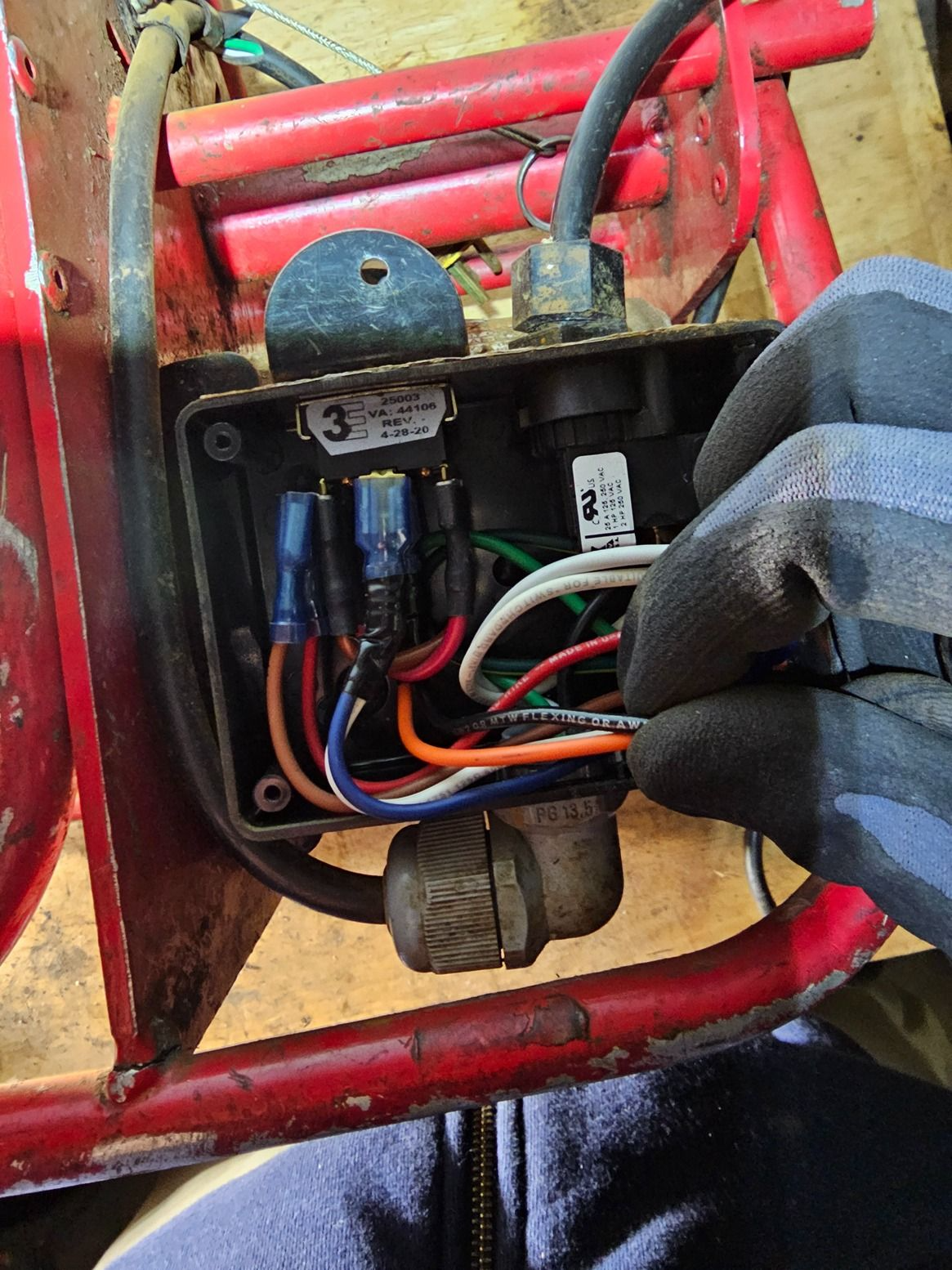 Close-up of a black electrical control box with wires mounted on a red metal frame, held by a gloved hand