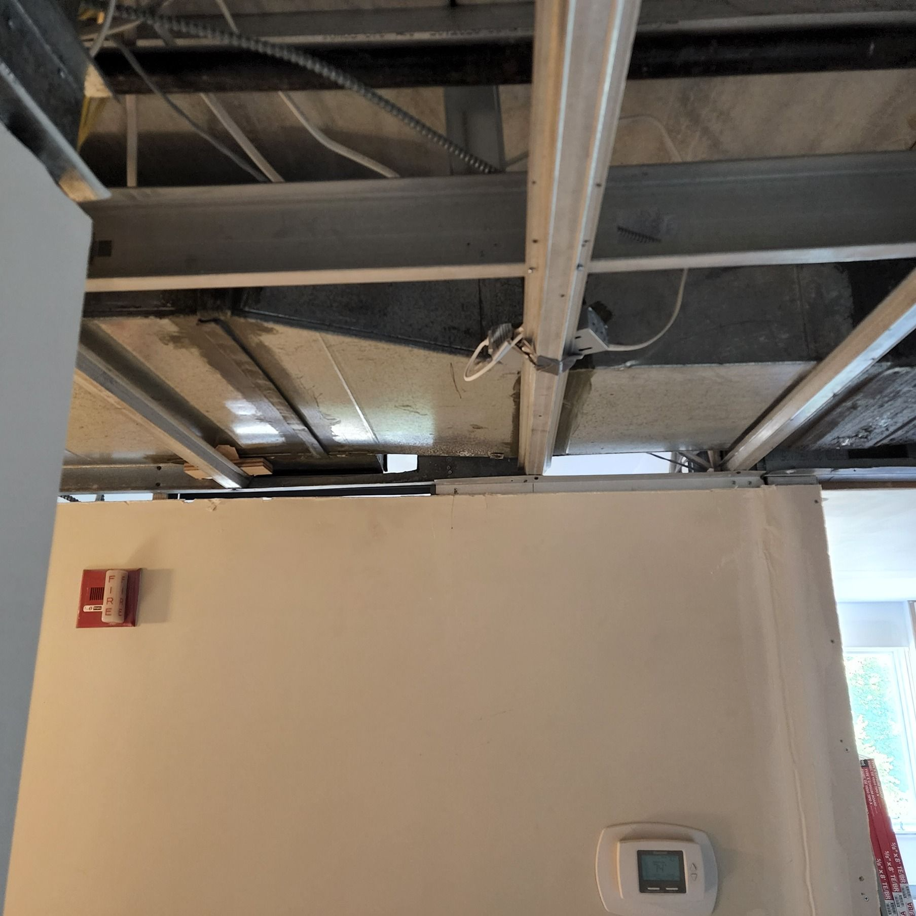 Indoor wall under exposed ceiling beams with hanging wires and a thermostat control panel.