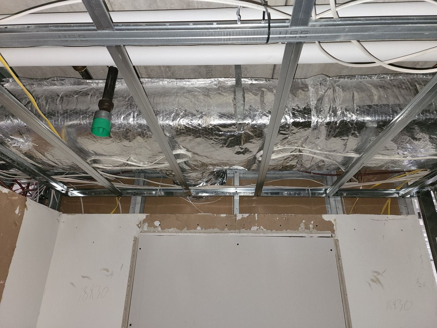 Exposed ceiling framing and ductwork with insulation above a white wall in a construction interior