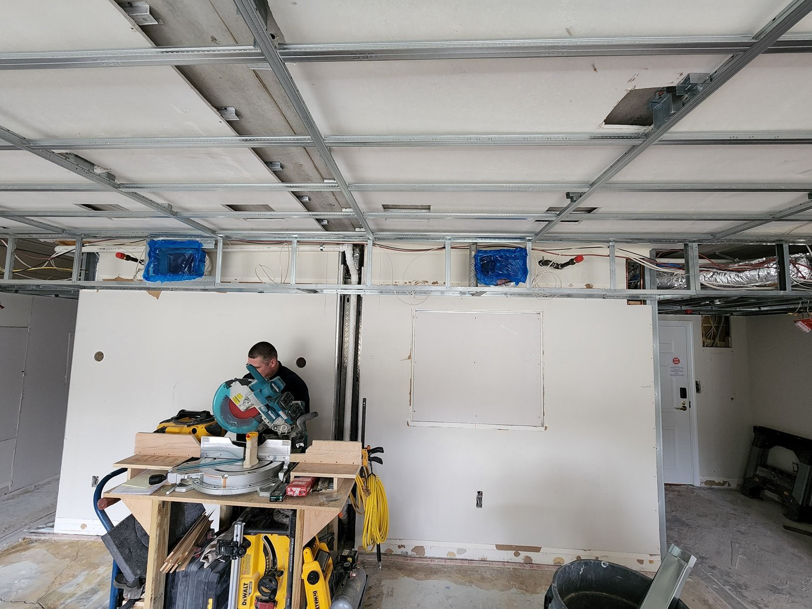 Worker using power tools in a white-walled room under construction with exposed ceiling framing