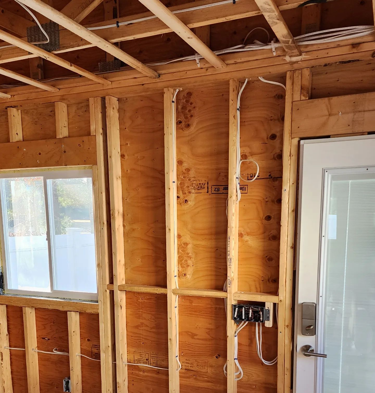 Exposed wooden wall framing and ceiling joists in a room, with a window and open electrical box.