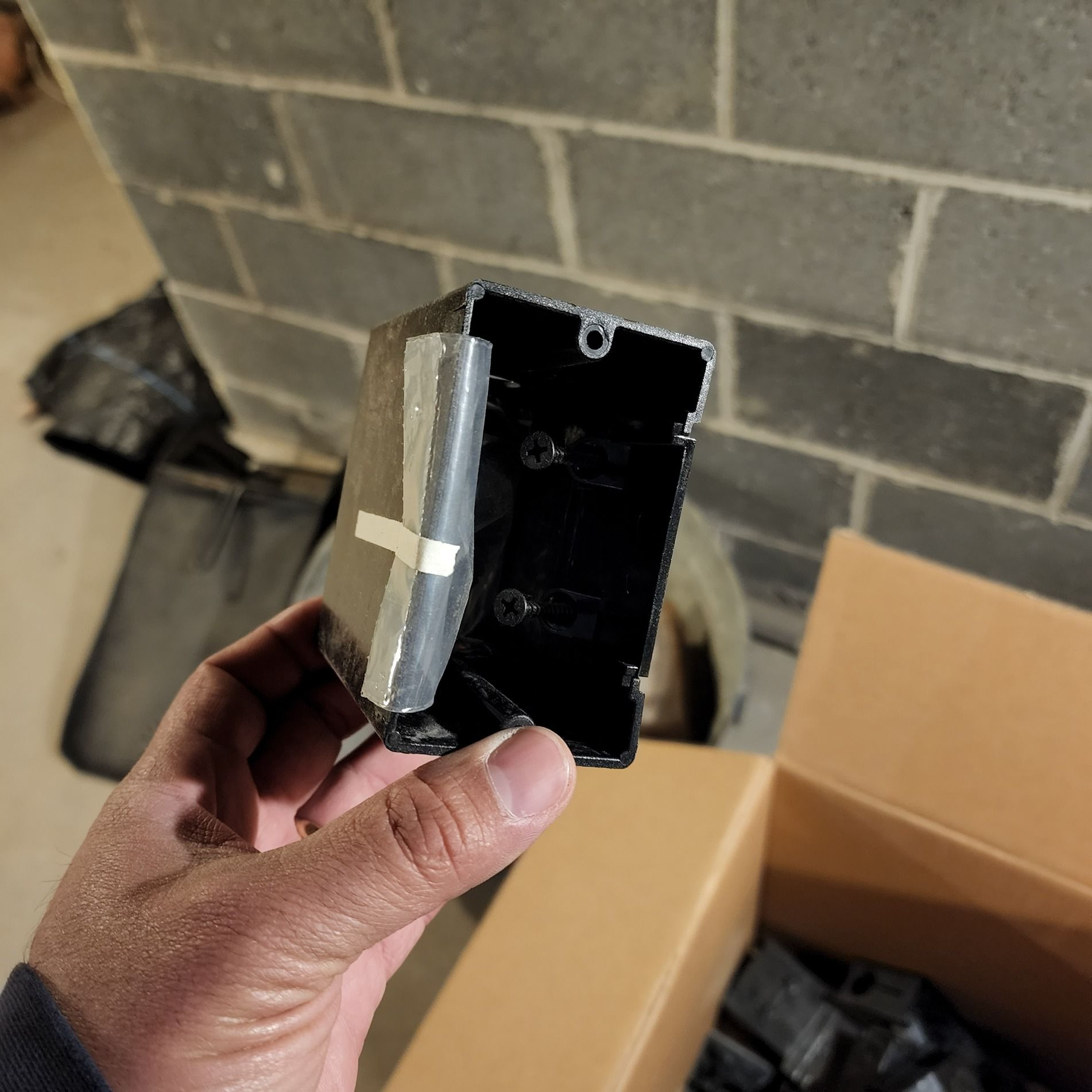 Hand holding a black electrical wall box with a taped silver bracket in front of open cardboard boxes