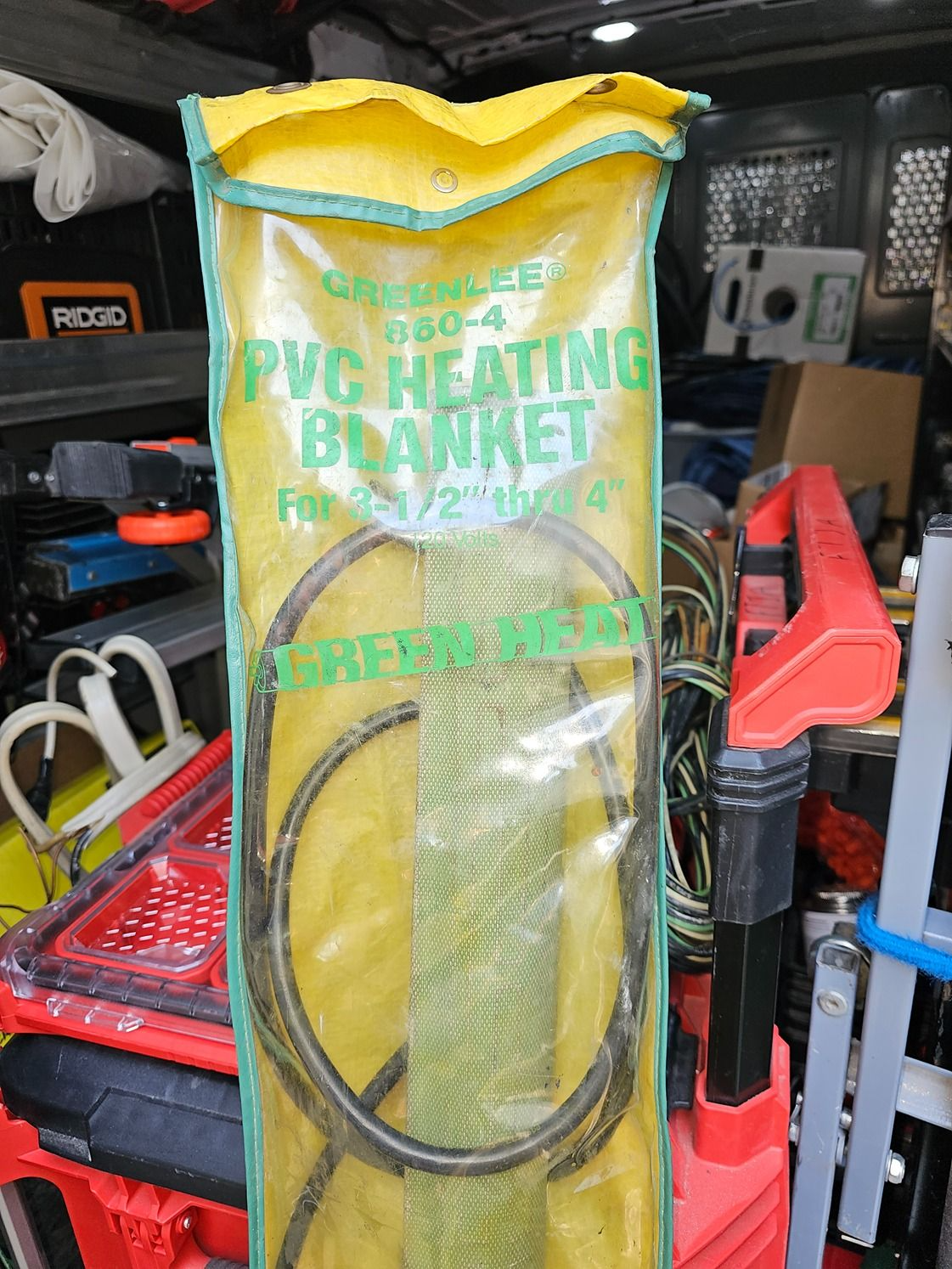 Yellow bag labeled “PVC heating blanket” standing in a cluttered workshop with tools and cables.