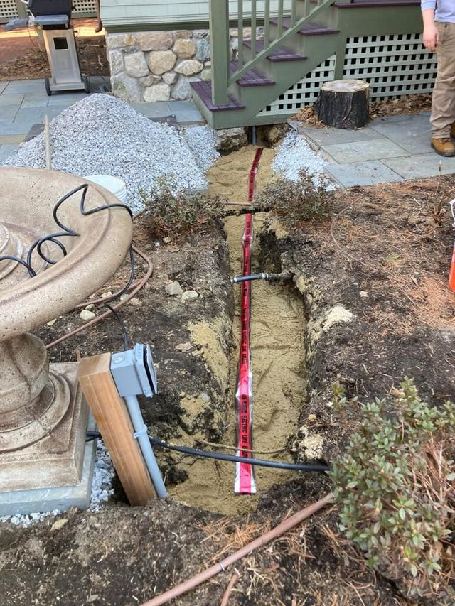 Yard trench with red conduit laid in dirt beside stones, steps, and a potted plant.