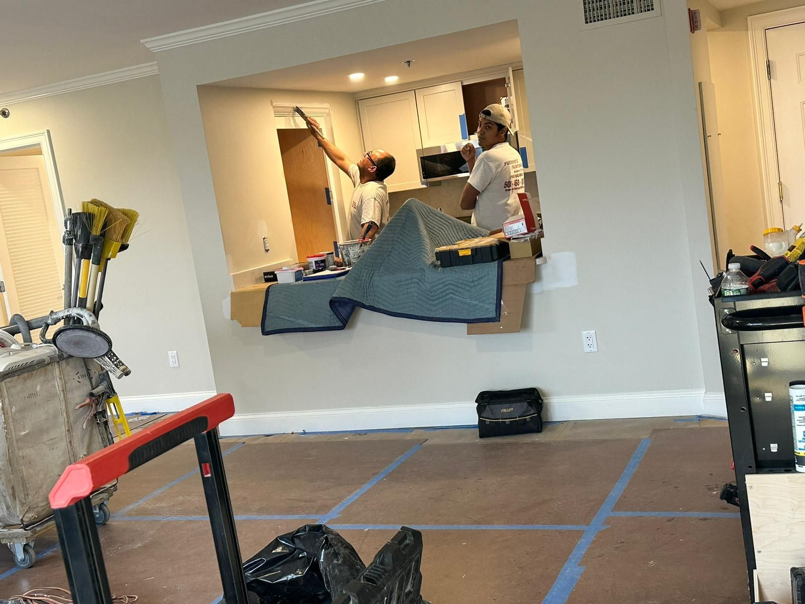 Workers renovating a room with blue taped floor, mirror opening, and tools on the left