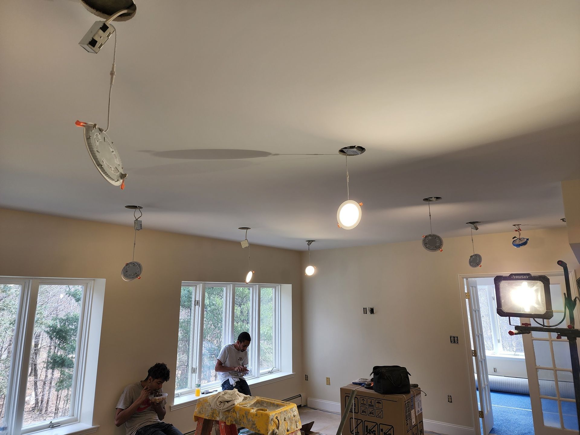 Bright living room with workers installing recessed lights and a ladder near large windows.