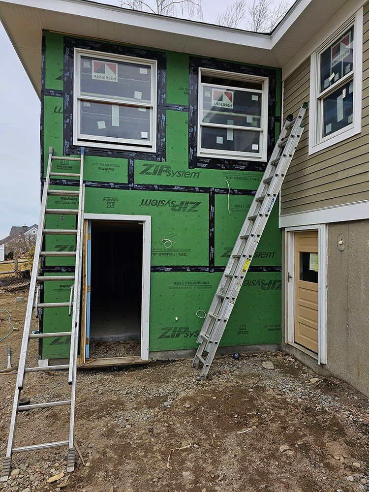 House exterior under renovation with green sheathing, two ladders, and newly installed windows over a doorway