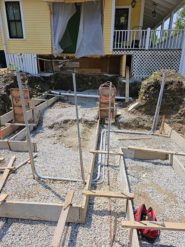 House foundation under construction with rebar, gravel, and formwork beneath a yellow porch