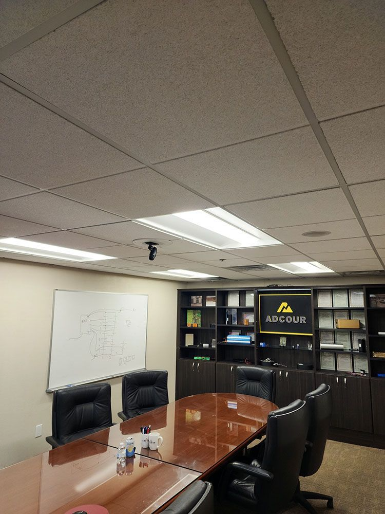 Conference room with a long wooden table, black chairs, whiteboard, and shelving under fluorescent lights.