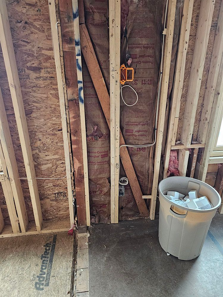 Exposed wall framing with insulation, electrical wiring, and a trash can on a concrete floor.