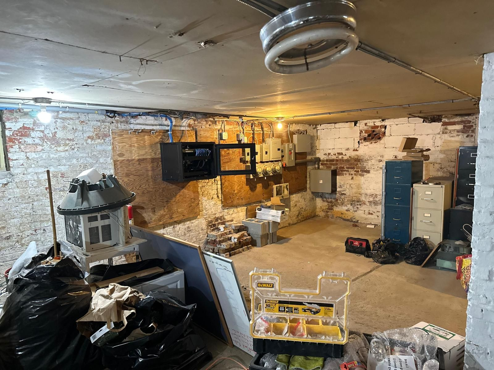 Cluttered unfinished basement with exposed brick, boxes, tools, and a hanging ceiling light.