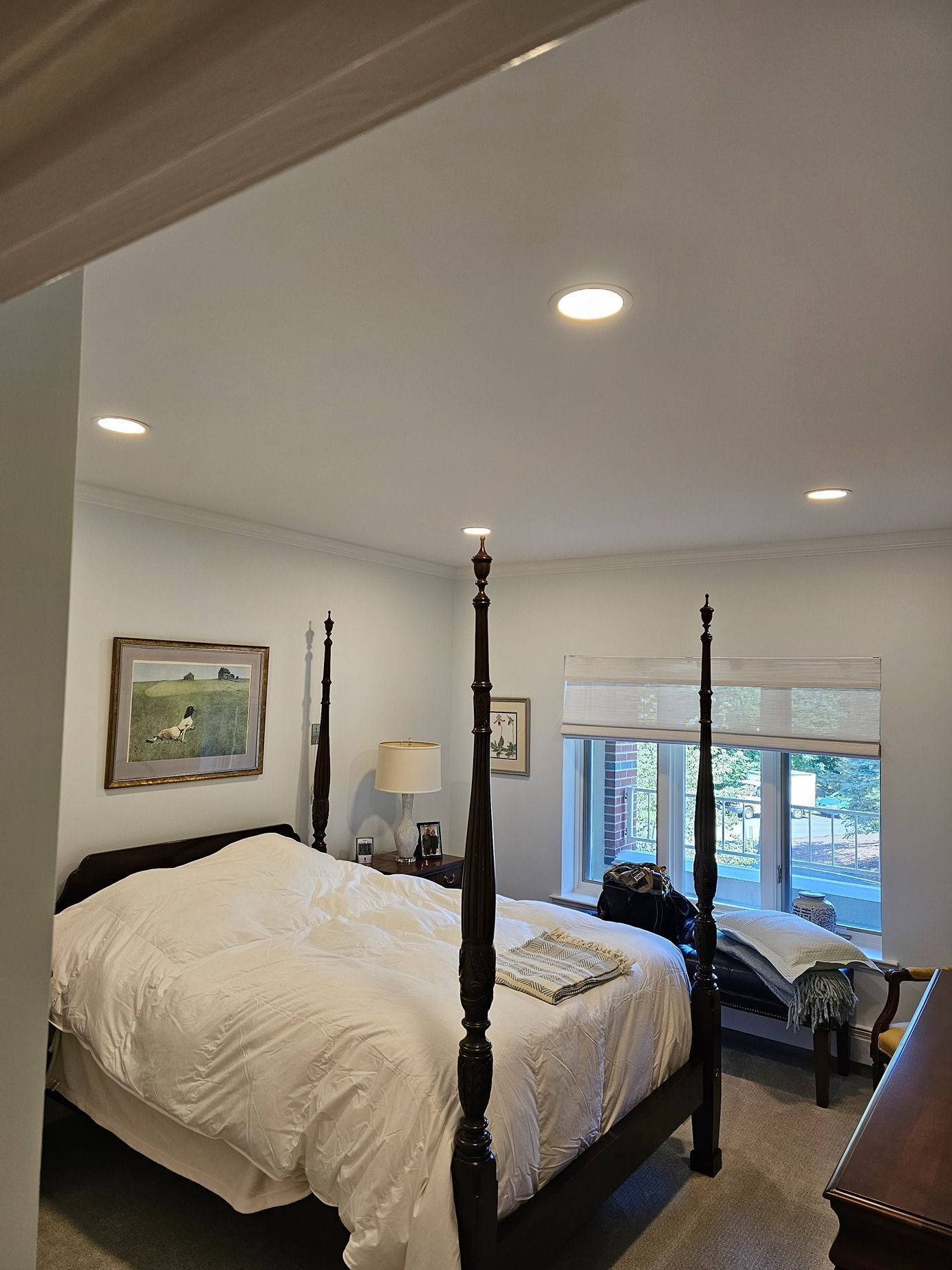 Bedroom with four-poster bed, white bedding, ceiling lights, and a window seat by a bright window.