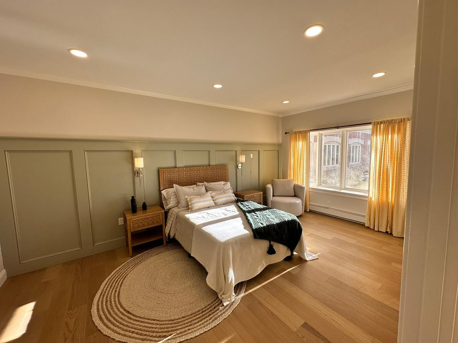 Bright bedroom with a bed, woven rug, wood floor, and sunlit windows with gold curtains.