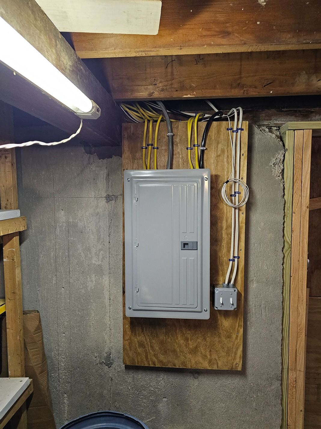 Electrical panel mounted on plywood in a basement with exposed wiring and wooden beams.