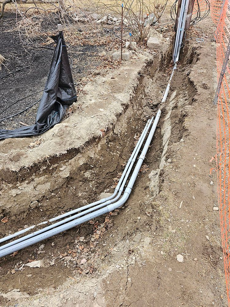 Trench with three white PVC pipes running along muddy ground beside orange construction netting