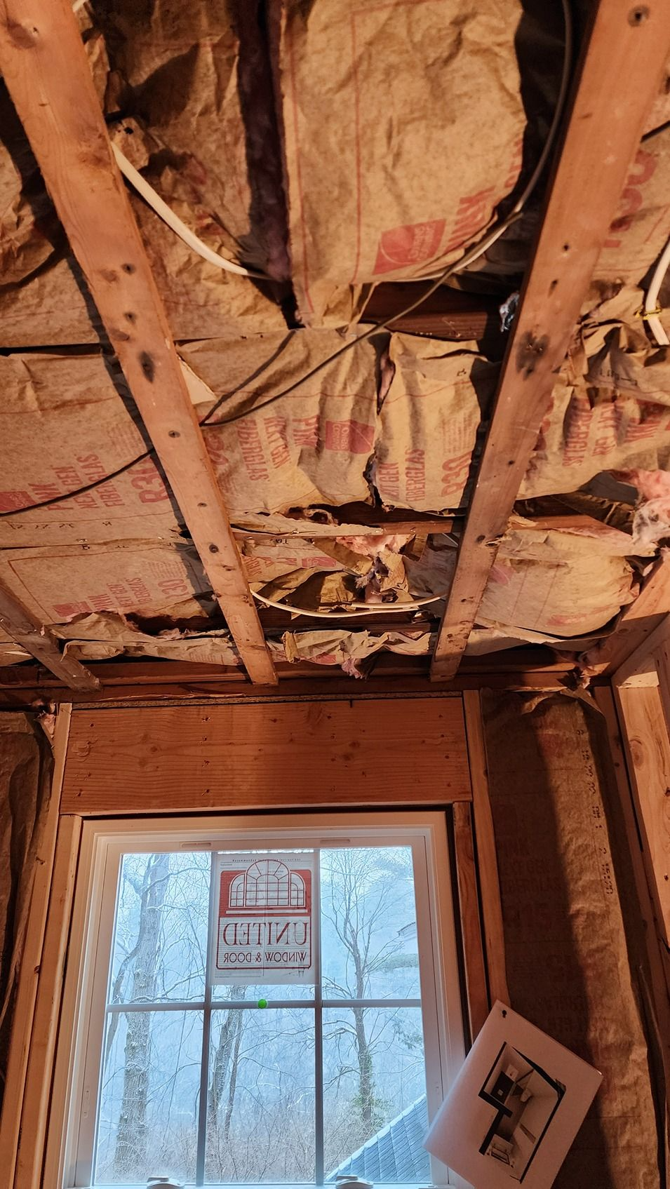 Partially demolished ceiling with exposed insulation and wiring above a window in a room under renovation