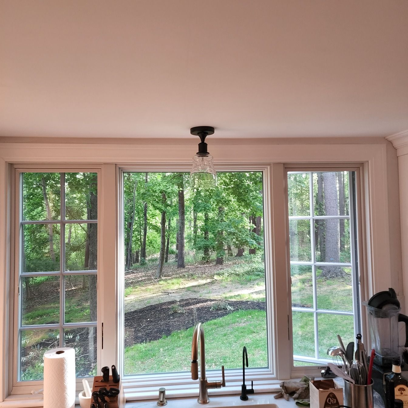 Kitchen window overlooking a green wooded yard with a sink and faucet in the foreground