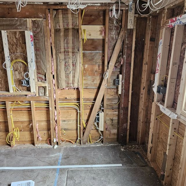 Exposed wooden wall studs and wiring in a room under renovation, with unfinished floor and ceiling joists.