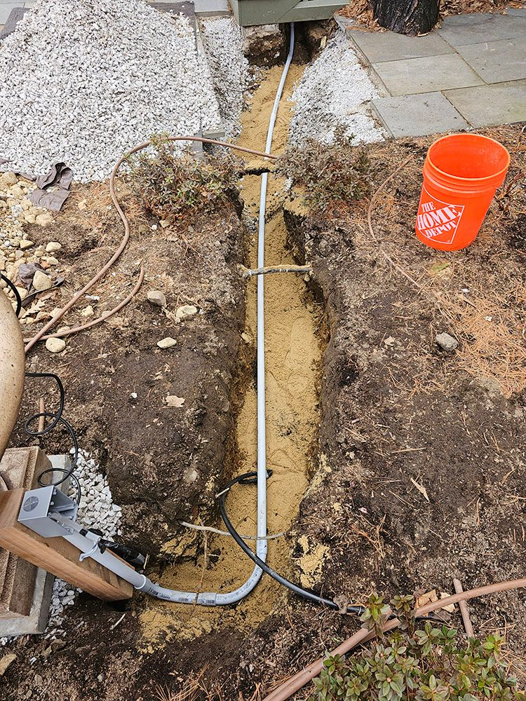 Trench dug through soil beside a gravel path, with a hose or cable running along the center and an orange bucket nearby