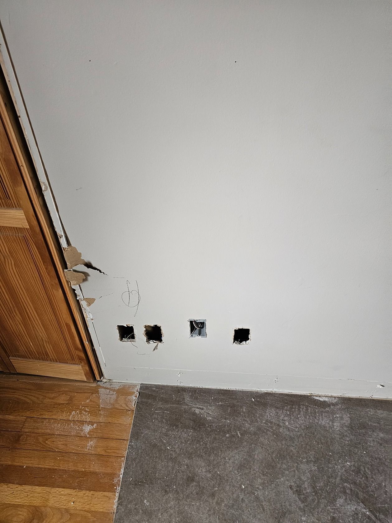 Damaged white wall beside a wooden door frame, with several open cable holes near the floor and carpet below