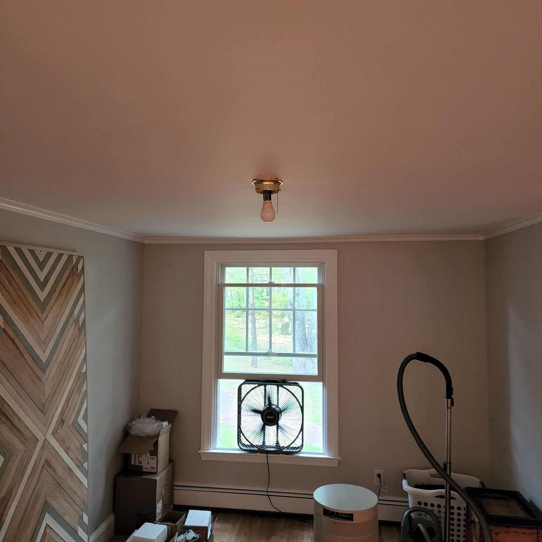 Small room with gray walls, a window fan, bare bulb ceiling light, and a vacuum cleaner near the right wall