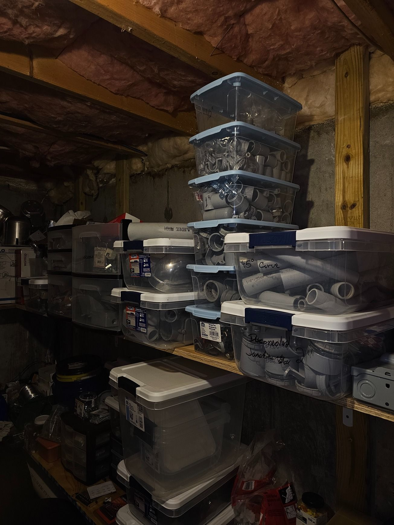 Stacked storage bins and plastic containers on shelves in a dim basement or workshop.