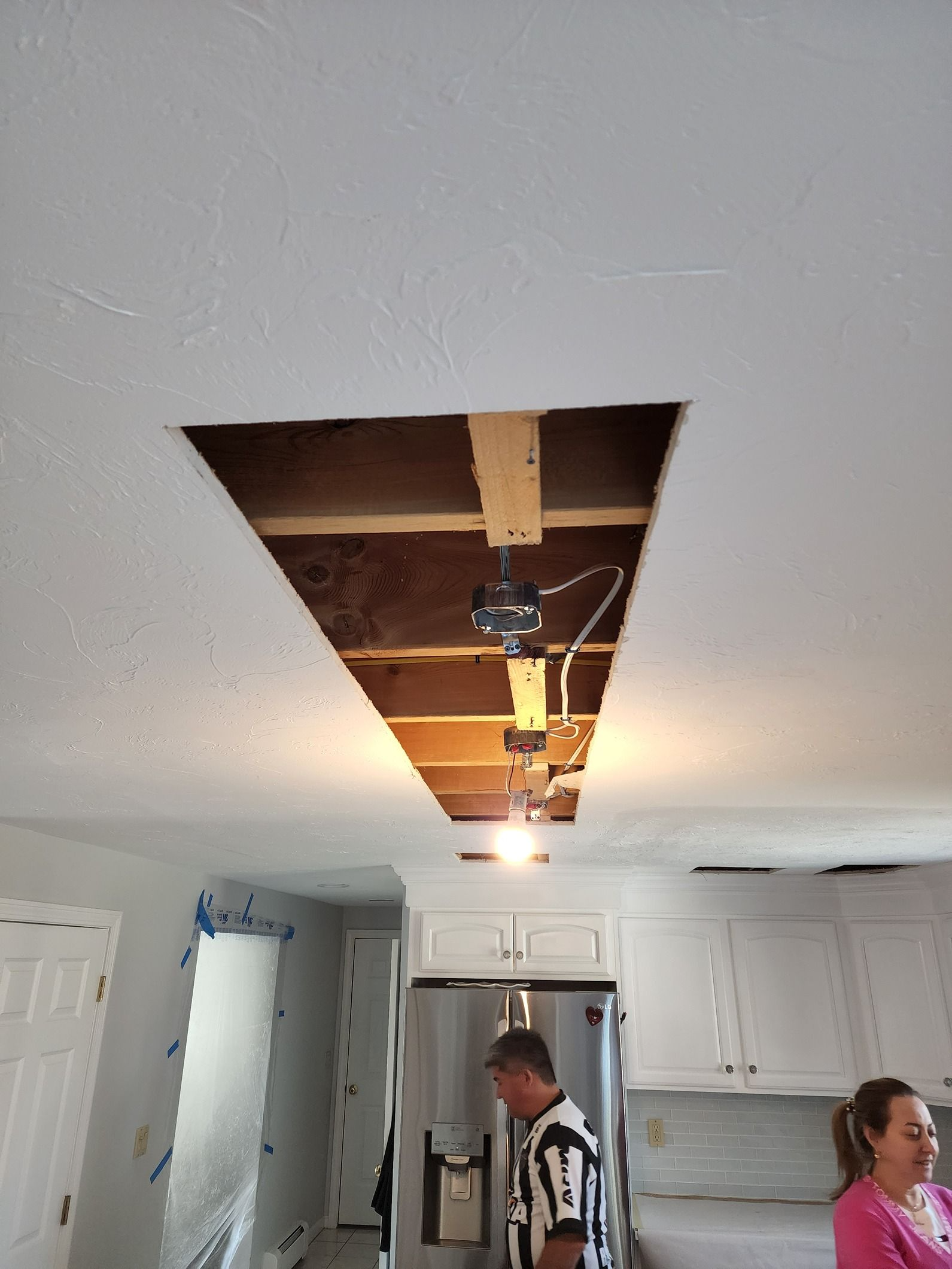 Kitchen ceiling under repair with a large drywall cutout exposing pipes and lights; two people standing below