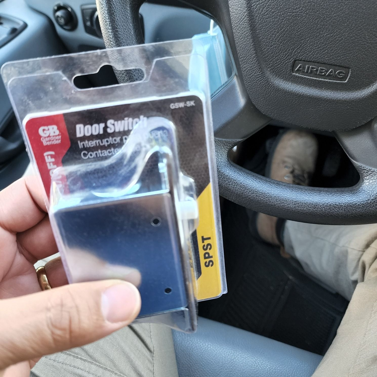 Hand holding a packaged black door switch in a car interior, with steering wheel visible.