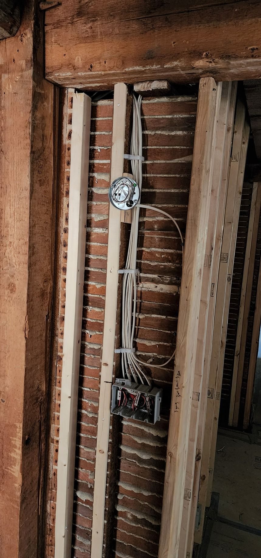 Exposed electrical wiring and outlets in an unfinished wall frame with wooden studs and brick backing