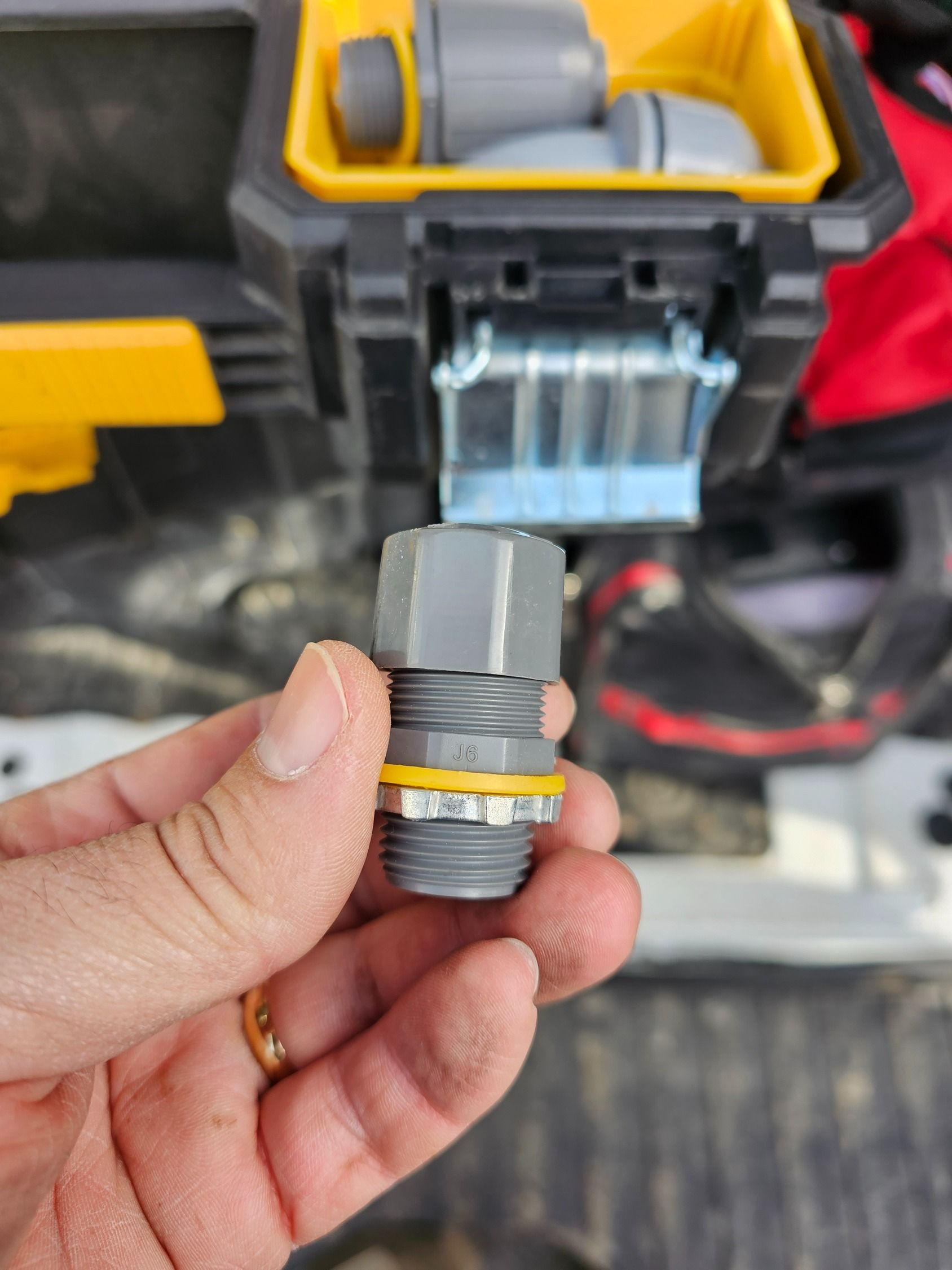 Hand holding a metal threaded fitting in front of a yellow-and-black toolbox battery compartment
