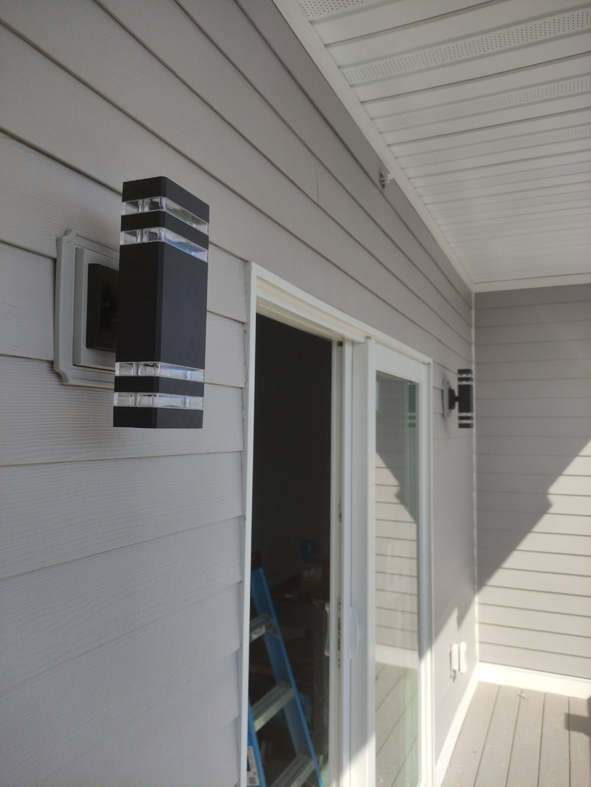 Exterior walkway with gray siding, wall lights, and an open doorway revealing a ladder inside.