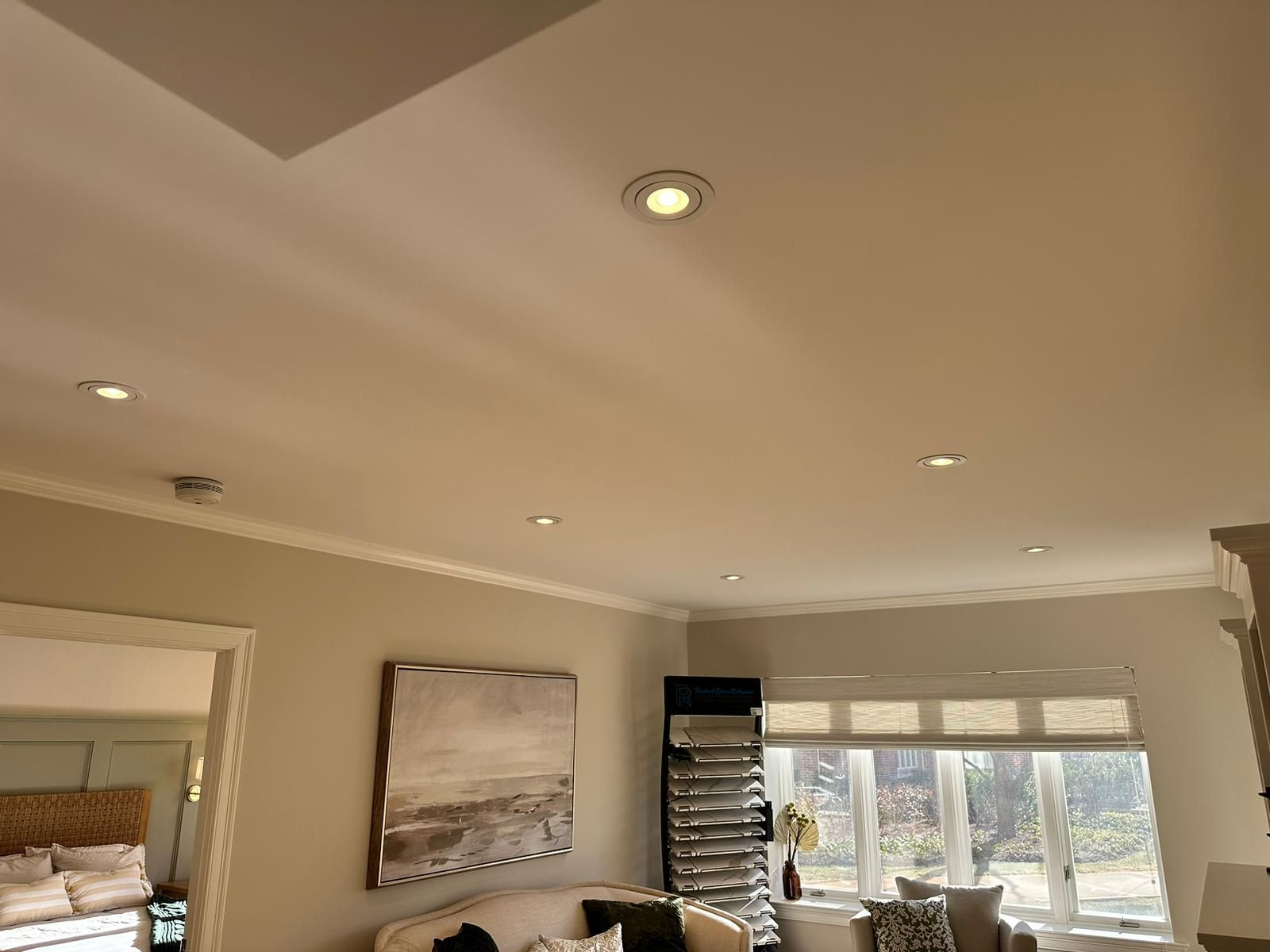Bright living room with recessed ceiling lights, wall art, and large windows over a cushioned chair.