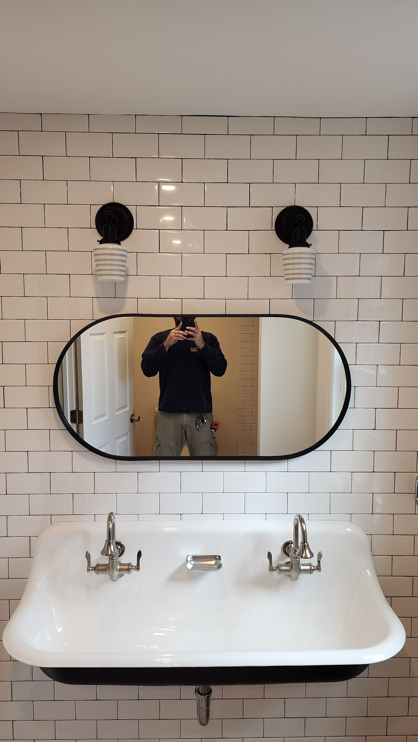 Bathroom sink and oval mirror on a white tiled wall, with a person taking the photo reflected in the mirror.