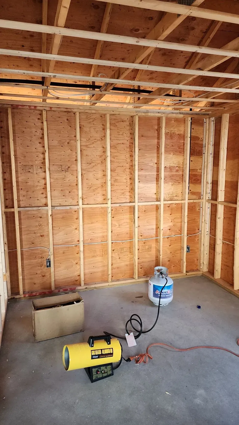 Unfinished wooden room with exposed studs, concrete floor, and a yellow space heater on the floor.