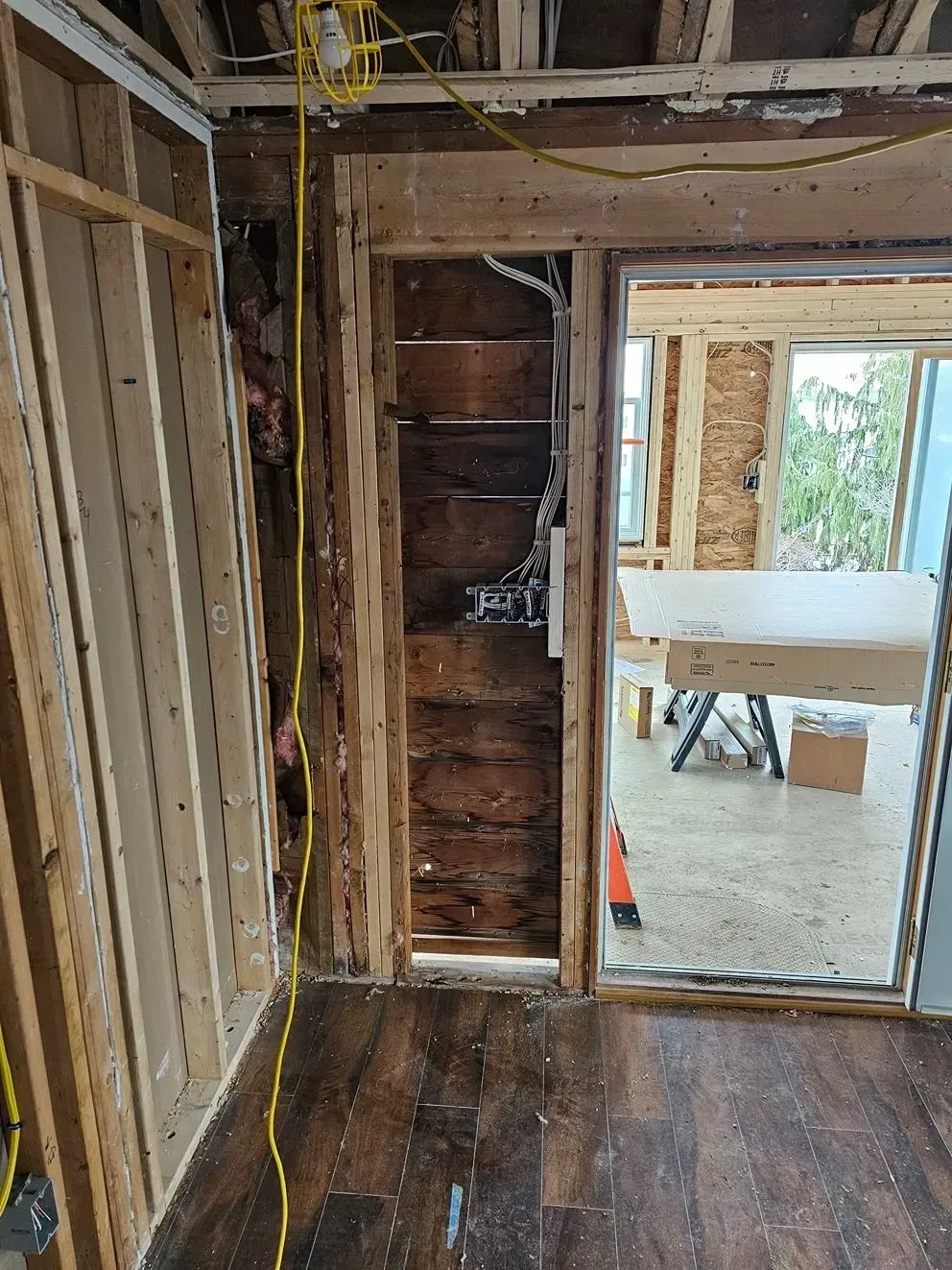 Partially gutted room under renovation, with exposed studs, open wall cavity, and a doorway to a brighter adjacent room