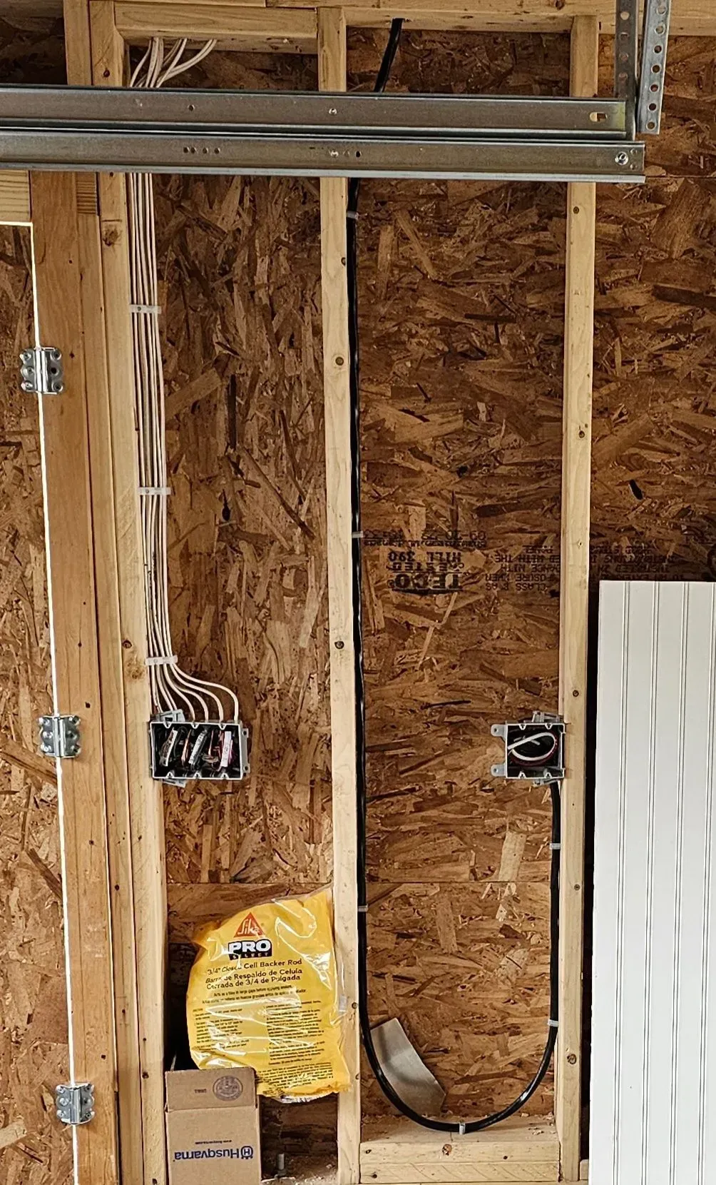Unfinished garage wall with exposed studs, wiring, outlets, insulation, and a yellow bag on the floor